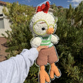 A handmade crocheted chicken doll being held up by a person's hand outdoors in front of green bushes and a blue sky. The doll has white yarn feathers, an orange beak and feet, a green striped shirt, brown shorts, and a red comb.