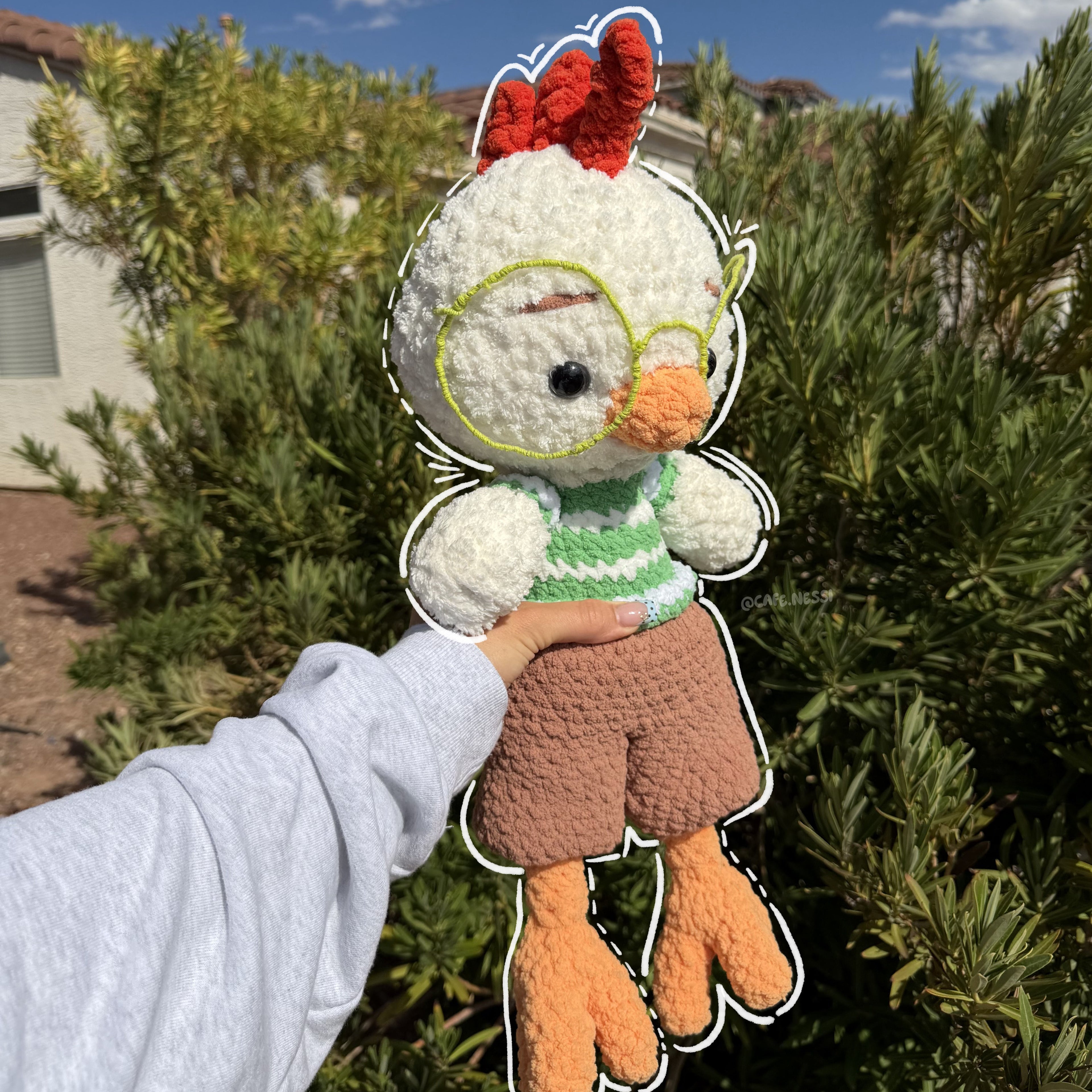 A handmade crocheted chicken doll being held up by a person's hand outdoors in front of green bushes and a blue sky. The doll has white yarn feathers, an orange beak and feet, a green striped shirt, brown shorts, and a red comb.