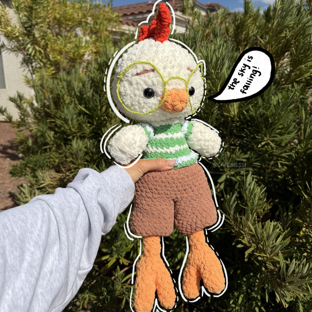 A handmade crochet chicken plush toy being held up by a person's hand outdoors. The plush has white feathers, a red comb, yellow-rimmed glasses, a green and white striped shirt, brown shorts, and orange legs.