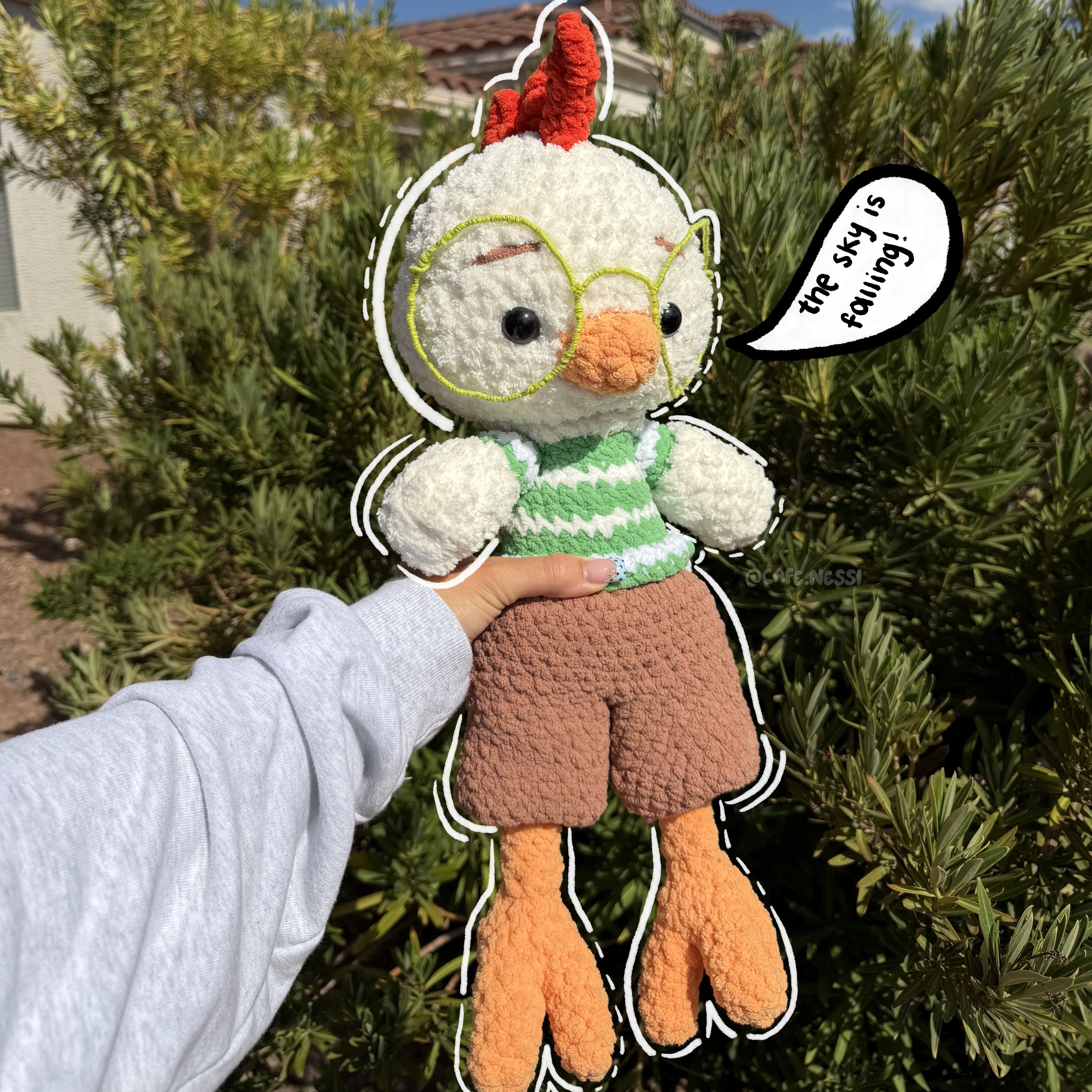 A handmade crochet chicken plush toy being held up by a person's hand outdoors. The plush has white feathers, a red comb, yellow-rimmed glasses, a green and white striped shirt, brown shorts, and orange legs.