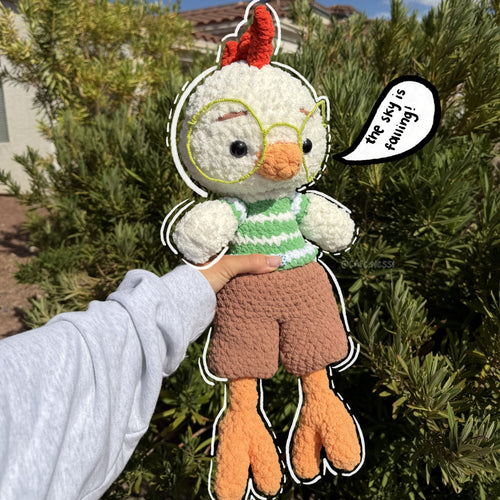 A handmade crochet chicken plush toy being held up by a person's hand outdoors. The plush has white feathers, a red comb, yellow-rimmed glasses, a green and white striped shirt, brown shorts, and orange legs.