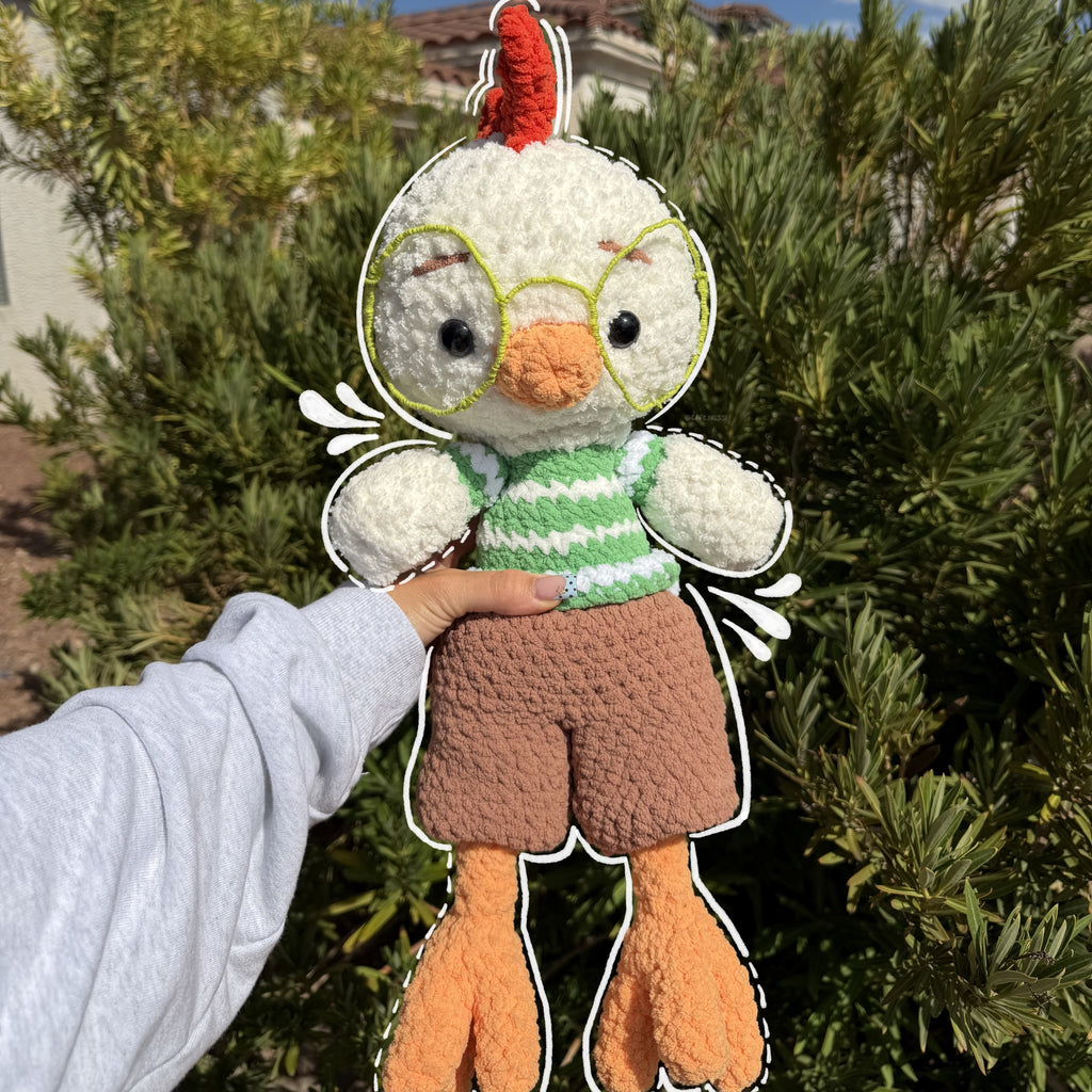 A handmade crocheted chick doll with a red comb, green striped sweater, brown shorts, and orange feet, held up by a person's hand against a background of green bushes and a blue sky.